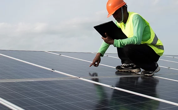 Man wearing a safety vest holding a tablet squatting down over solar panels